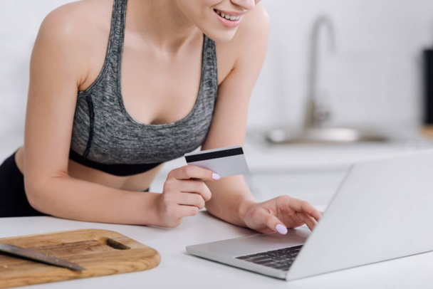 cropped view of cheerful girl holding credit card near laptop in kitchen   - Photo, Image