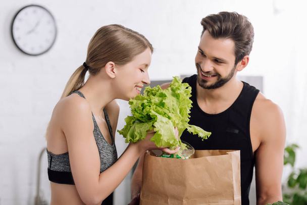 cheerful girl holding lettuce near paper bag and happy bearded man in kitchen  - Photo, Image