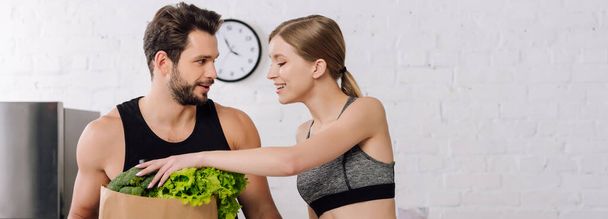 panoramic shot of cheerful girl taking lettuce from paper bag near bearded man in kitchen  - Photo, Image