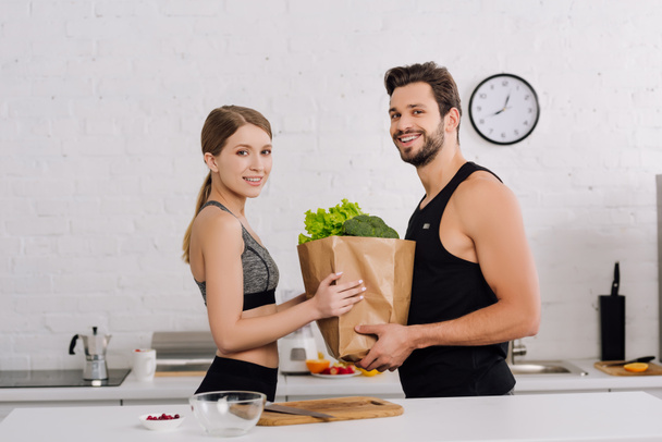 счастливая девушка и бородатый мужчина держит бумажный пакет с продуктами на кухне
  - Фото, изображение