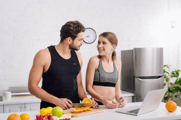 happy girl looking at sportive man near laptop and fruits in kitchen  - Фото, изображение