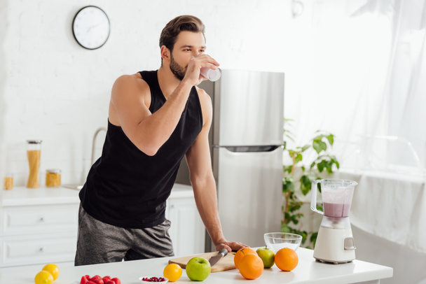 brodaty mężczyzna pijący świeży koktajl w pobliżu blendera i owoców  - Zdjęcie, obraz