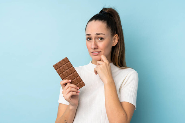 Young brunette woman over isolated blue background taking a chocolate tablet and having doubts - 写真・画像