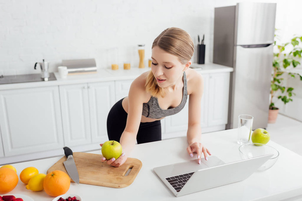 happy and sportive woman looking at tasty apple near laptop  - Foto, Imagem