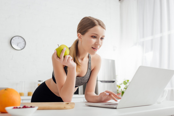 selective focus of sportive woman holding tasty apple and using laptop  - Fotoğraf, Görsel