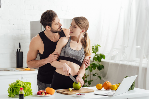 boldog lány vágás alma közelében sportos férfi, laptop és gyümölcsök a konyhában  - Fotó, kép