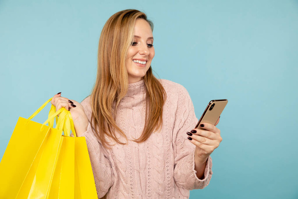 Młoda uśmiechnięta kobieta uśmiecha się i używa telefonu trzymając żółte torby na zakupy odizolowane. - Zdjęcie, obraz