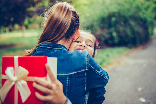 Two school friends hugging, emotional meeting in the park. Happy little girl gives her smiling friend a gift in a red box, making a surprise. People, children, holidays, friends and friendship concept - Fotó, kép