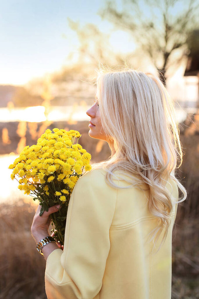 schöne Blondine im gelben Mantel und mit einem Strauß gelber Chrysanthemen steht am Fluss - Foto, Bild