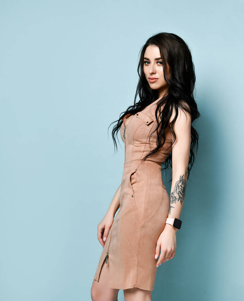 Brunette model in beige dress. There is watch on her tattooed hand. Standing sideways against blue studio background. Close up - Foto, afbeelding