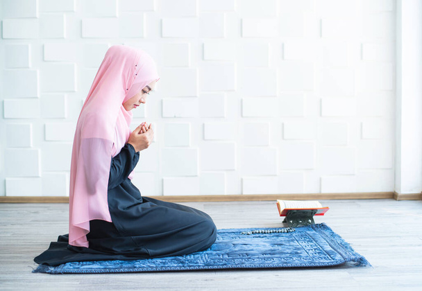 vista lateral de asiático hermosa joven musulmana estudiante oración en hijab orando en alfombra alfombra interior
 - Foto, Imagen