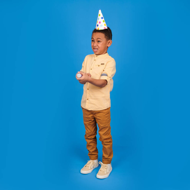 Excited Afro-American boy popping cracker and closing eyes. Playful little boy in white t-shirt and hat exploding party popper standing on blue studio background - Photo, image