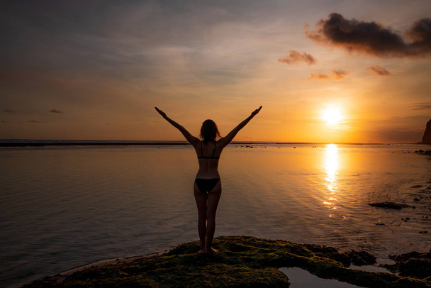 Travel lifestyle. Excited young woman raising arms at the beach in front of the ocean. View from back. Sunset golden hour at the beach. Amazing scenery. Sun reflection in water. Melasti beach, Bali - Photo, Image