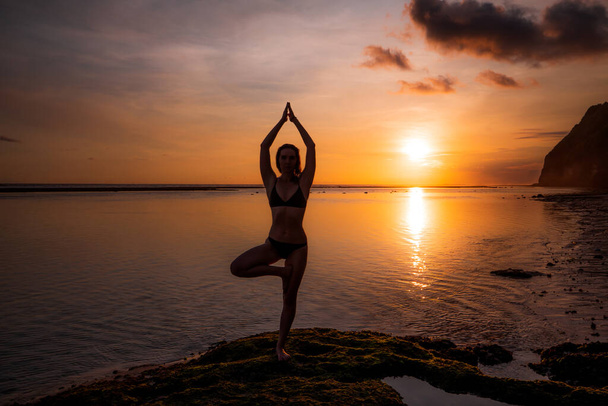 Vrikshasana asana 。日没の黄金の時間の間にビーチで木のポーズを練習若い女性。ナマストムドラで腕を上げる。屋外ヨガ。水の中の太陽の反射。バリ島のメラスティビーチ - 写真・画像