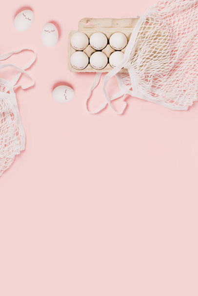 Happy Easter. White eggs with painted eyes in paper packaging and white mesh bag on pink background. Flat lay, top view, copy space. - Foto, afbeelding