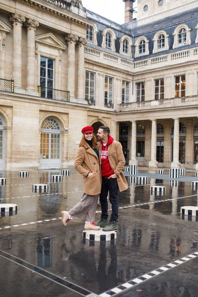 A young beautiful couple is stylishly dressed, a girl in a brown coat and a red hat, a man in a brown coat and red sweater are standing in a square in Paris France - Фото, изображение