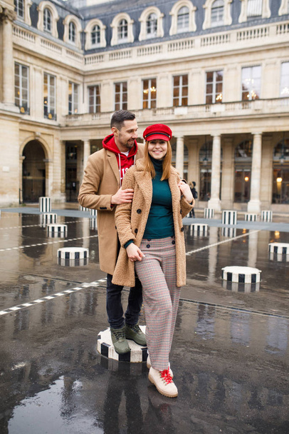 A young beautiful couple is stylishly dressed, a girl in a brown coat and a red hat, a man in a brown coat and red sweater are standing in a square in Paris France - Foto, afbeelding