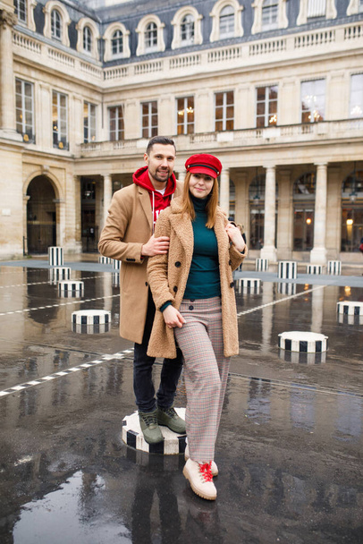 A young beautiful couple is stylishly dressed, a girl in a brown coat and a red hat, a man in a brown coat and red sweater are standing in a square in Paris France - Photo, image