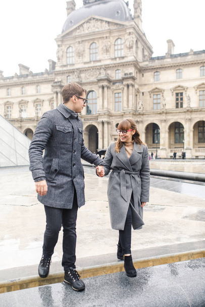 Young beautiful couple dressed stylishly, a girl in a gray coat, a man in a gray coat are walking near the Louvre Museum in Paris France - Photo, Image