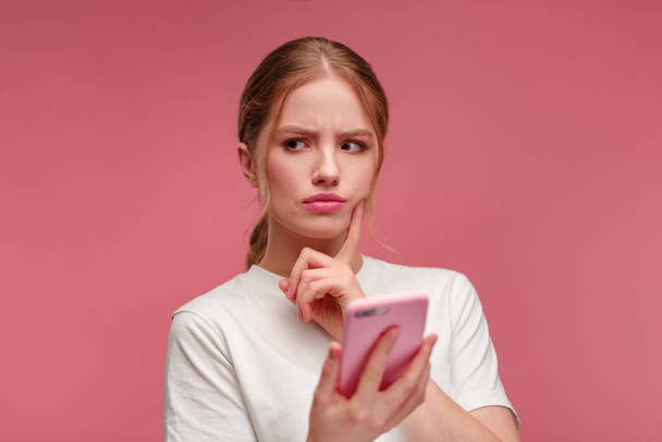 Confused young redhead woman holding pink smartphone.Embarassed girl has problem with broken not working mobile phone.Receiving bad news or email,spam message.Young people working with mobile devices - Фото, изображение