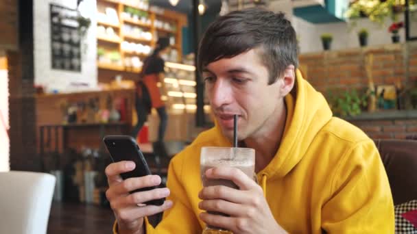 Mann mit Smartphone-Touchscreen in modernem Café. Männchen trinken Milchshake oder Smoothie und SMS auf Smartphone im Café - Filmmaterial, Video