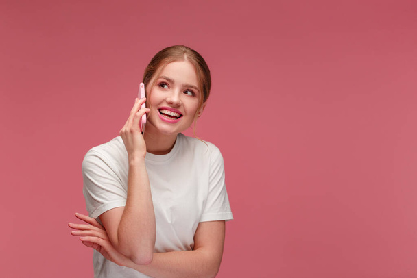 Portrait of cute happy redhead girl wearing white t-shirt talking on mobile phone and smiling isolated over pink background. Copy space. Young people working with mobile devices - Photo, image