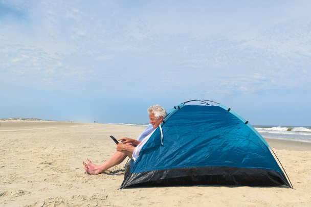 Single senior man with digital tablet camping in shelter at the beach - Foto, afbeelding