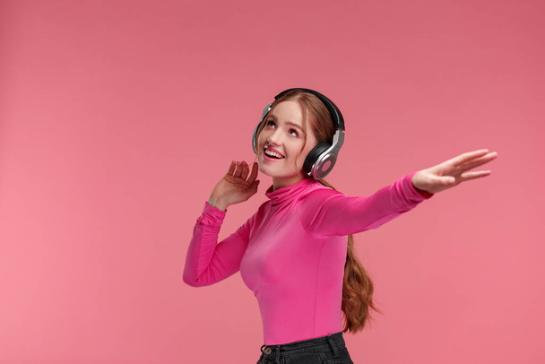 Enjoy listening to music. Beautiful young redhead woman in headphones listening music. Funny smiling girl in earphones and pink blouse dancing on pink background. Relaxation and stress management - Foto, immagini