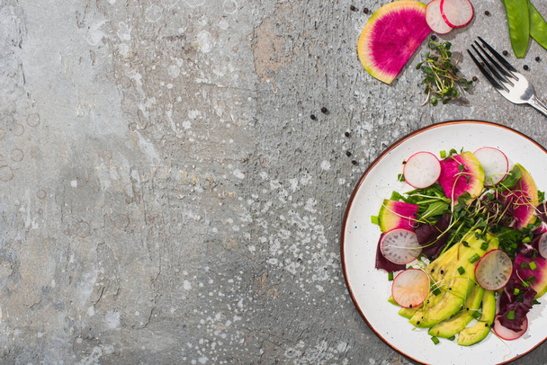 top view of fresh radish salad with greens and avocado on grey concrete surface with fork - Foto, Imagen