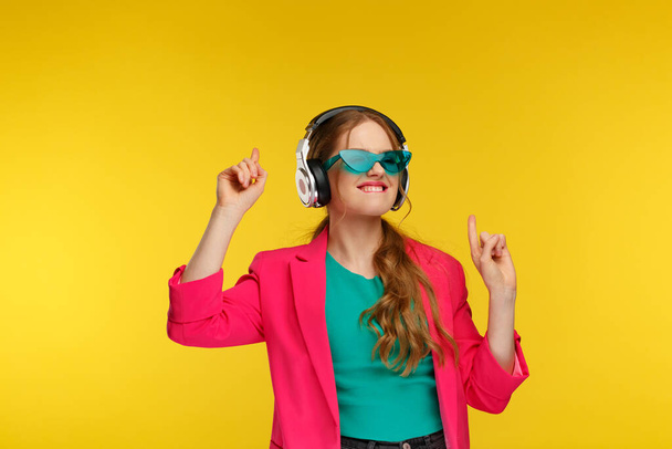 Enjoy listening to music. Young redhead woman in headphones listening music. Funny smiling girl in earphones and pink jacket dancing and singing on yellow background. Relaxation and stress management - 写真・画像