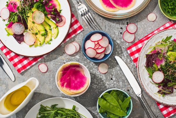 top view of fresh radish salad with greens and avocado served on plates on grey concrete surface with ingredients, cutlery and napkins - Photo, Image