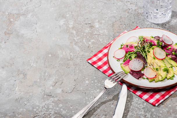 fresh radish salad with greens and avocado served on napkin with cutlery near water on grey concrete surface - Φωτογραφία, εικόνα
