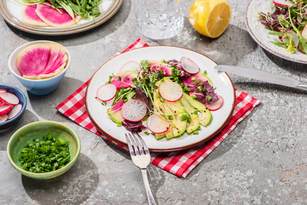 fresh radish salad with greens and avocado on napkin on grey concrete surface with ingredients in bowls and cutlery - Photo, Image