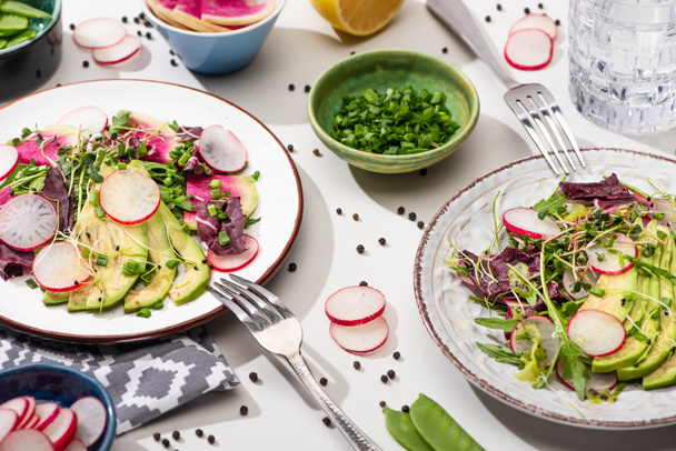 fresh radish salad with greens and avocado on plates on white surface with ingredients in bowls and water - Photo, Image