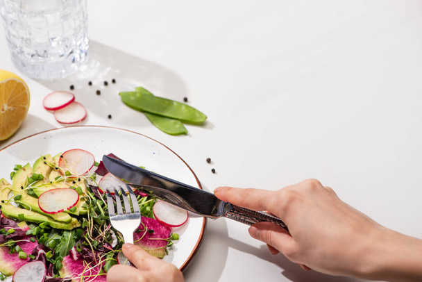 vista ritagliata di donna che mangia insalata di ravanello fresco con verdure e avocado su superficie bianca con acqua
 - Foto, immagini