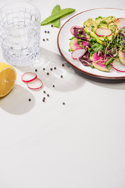 свіжий салат з редькою з зеленню та авокадо на тарілці на білій поверхні з водою, лимон
 - Фото, зображення