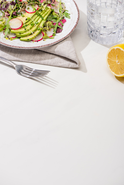 свіжий салат з редькою з зеленню та авокадо на тарілці на білій поверхні з лимоном та водою
 - Фото, зображення