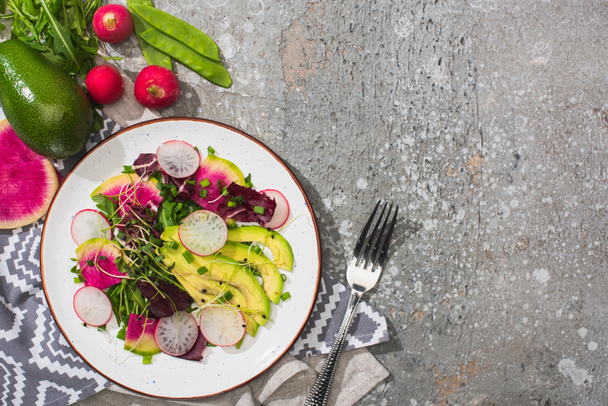 top view of fresh radish salad with greens and avocado near fork and vegetables on grey concrete surface - Photo, Image
