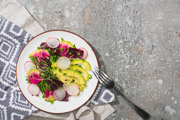 top view of fresh radish salad with greens and avocado near fork on grey concrete surface with napkins - Photo, Image