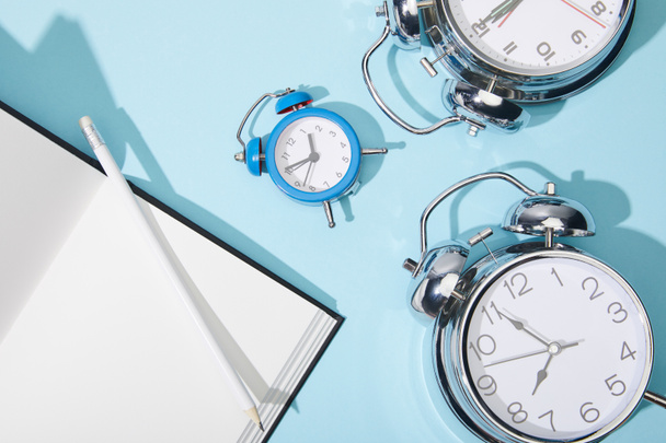 top view of classic alarm clocks with blank netbook and pencil on blue background - Foto, afbeelding
