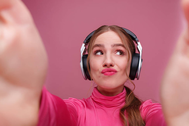 Gros plan portrait d'une jeune femme rousse souriante faisant du selfie à deux mains. Funny fille souriante portant des écouteurs donnant un cadeau isolé sur fond rose. Espace de copie - Photo, image