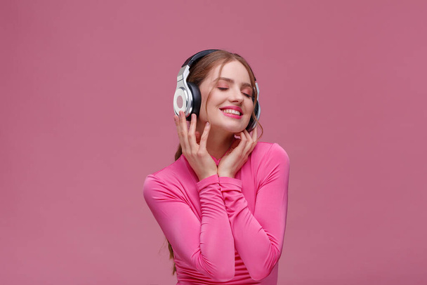 Enjoy listening to music. Beautiful young redhead woman in headphones listening music. Funny smiling girl in earphones and pink blouse dancing on pink background. Relaxation and stress management - Fotoğraf, Görsel