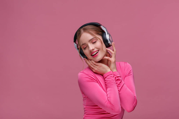 Enjoy listening to music. Beautiful young redhead woman in headphones listening music. Funny smiling girl in earphones and pink blouse dancing on pink background. Relaxation and stress management - Foto, afbeelding