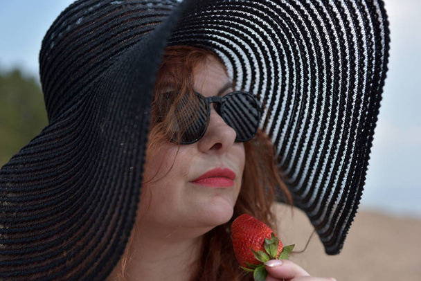 portrait of a woman in a wide-brimmed hat and sunglasses with strawberries in her hands - Фото, изображение
