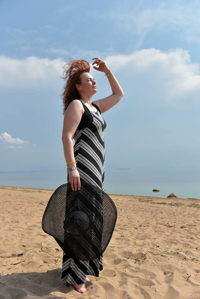 woman in a striped dress on a background of the sea with a hat in her hands - Photo, image