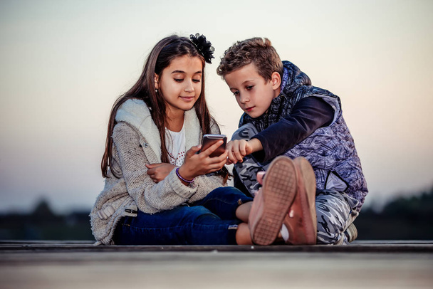 Δύο νεαροί χαριτωμένοι μικροί φίλοι, αγόρι και κορίτσι που βλέπουν κάτι στο smartphone να κάθεται δίπλα στη λίμνη το βράδυ. Τα παιδιά περνάνε καλά μαζί. Φιλία - Φωτογραφία, εικόνα