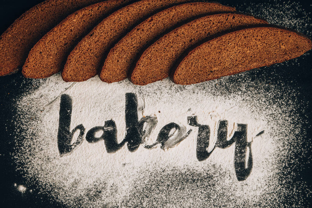 Aufschrift Bäckerei auf weißem Weizenmehl verstreut in Scheiben geschnittenes Roggenbrot auf dunklem Hintergrund, handgeschrieben in Mehl das Wort Bäckerei - Foto, Bild