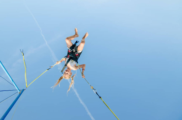 активная маленькая девочка-подросток, прыгающая на батуте высоко в голубое небо, защищенное эластичным ремнем безопасности - Фото, изображение
