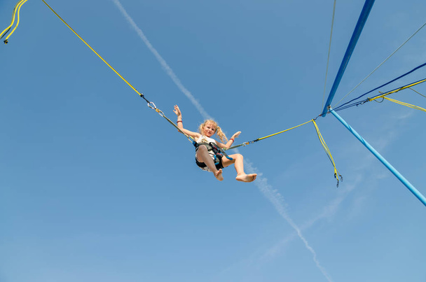 aktives kleines Teenie-Mädchen springt im Trampolin hoch in den blauen Himmel, gesichert mit elastischem Sicherheitsgurt - Foto, Bild