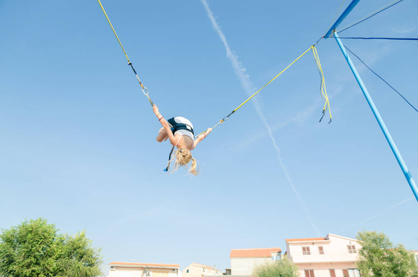 ενεργό μικρό κορίτσι έφηβος άλμα σε τραμπολίνο ψηλά στον γαλάζιο ουρανό με ελαστική ζώνη ασφαλείας - Φωτογραφία, εικόνα